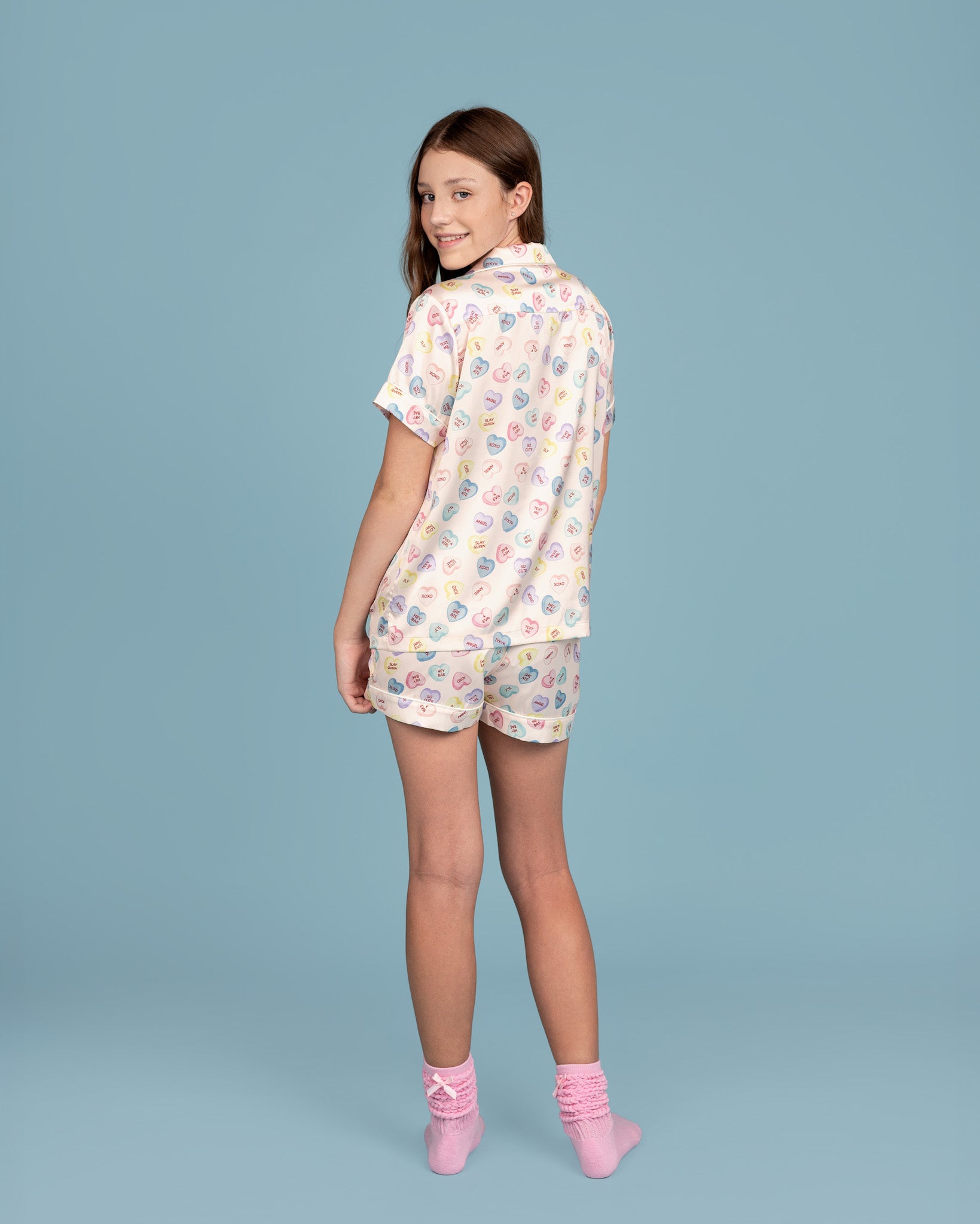 teen model wearing Dream Queen Co. Sleep Cute Short Sleeve satin pj set in conversation cutie pastel heart print, and pink socks